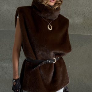 K&ouml;nnte beinhalten: Eine braune Kunstfellweste mit hohem Kragen und schwarzem G&uuml;rtel. Die Weste wird mit einer braunen Lederbarett und einer goldenen Anh&auml;ngerkette kombiniert. Das Outfit wird durch schwarze Lederhandschuhe erg&auml;nzt.