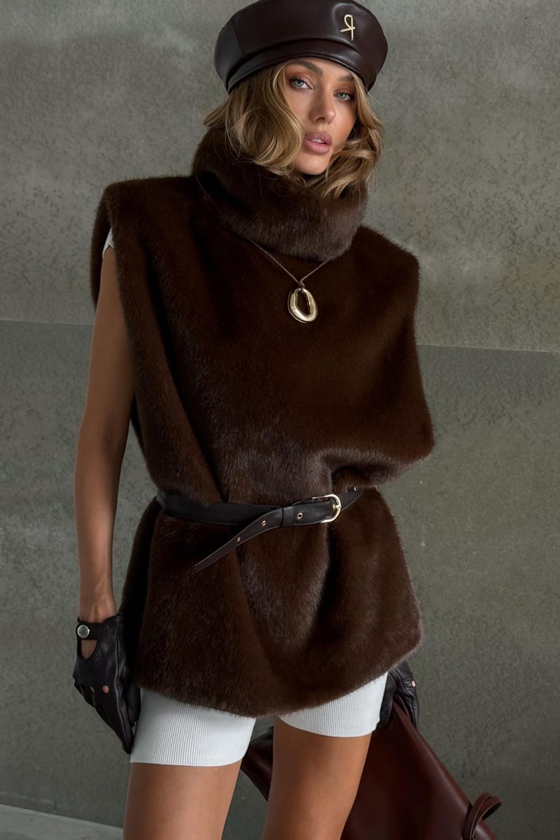 K&ouml;nnte beinhalten: Eine braune Kunstfellweste mit hohem Kragen und schwarzem G&uuml;rtel. Die Weste wird mit einer braunen Lederbarett und einer goldenen Anh&auml;ngerkette kombiniert. Das Outfit wird durch schwarze Lederhandschuhe erg&auml;nzt.