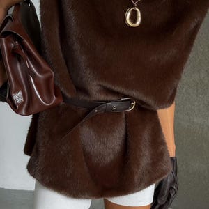 K&ouml;nnte beinhalten: Eine braune Kunstfellweste mit passendem G&uuml;rtel, kombiniert mit wei&szlig;en Shorts. Eine goldene Anh&auml;nger-Halskette und eine braune Ledertasche vervollst&auml;ndigen den Look. Das Outfit wird mit schwarzen Handschuhen gestylt.