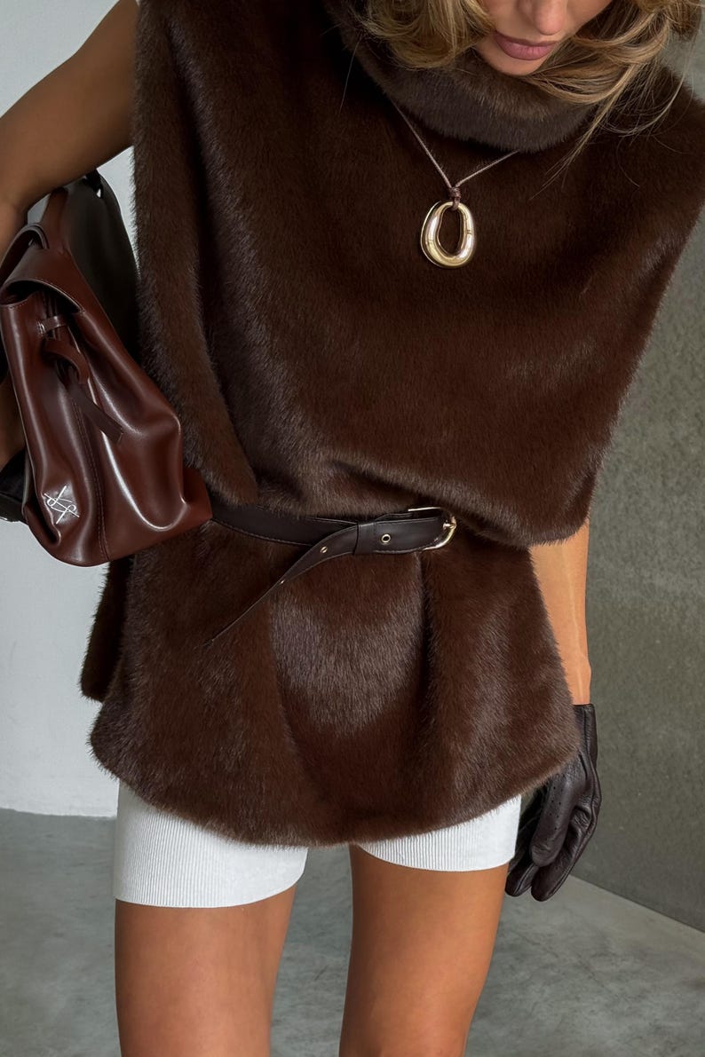 K&ouml;nnte beinhalten: Eine braune Kunstfellweste mit passendem G&uuml;rtel, kombiniert mit wei&szlig;en Shorts. Eine goldene Anh&auml;nger-Halskette und eine braune Ledertasche vervollst&auml;ndigen den Look. Das Outfit wird mit schwarzen Handschuhen gestylt.