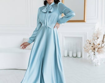 May include: A woman wearing a light blue satin dress with a bow at the neck. The dress has long sleeves and a flowing skirt. She is wearing nude heels.