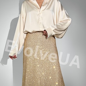 May include: A woman is wearing a long, gold sequined skirt with a cream colored satin shirt. The skirt has a subtle shimmer and is a great choice for a special occasion.