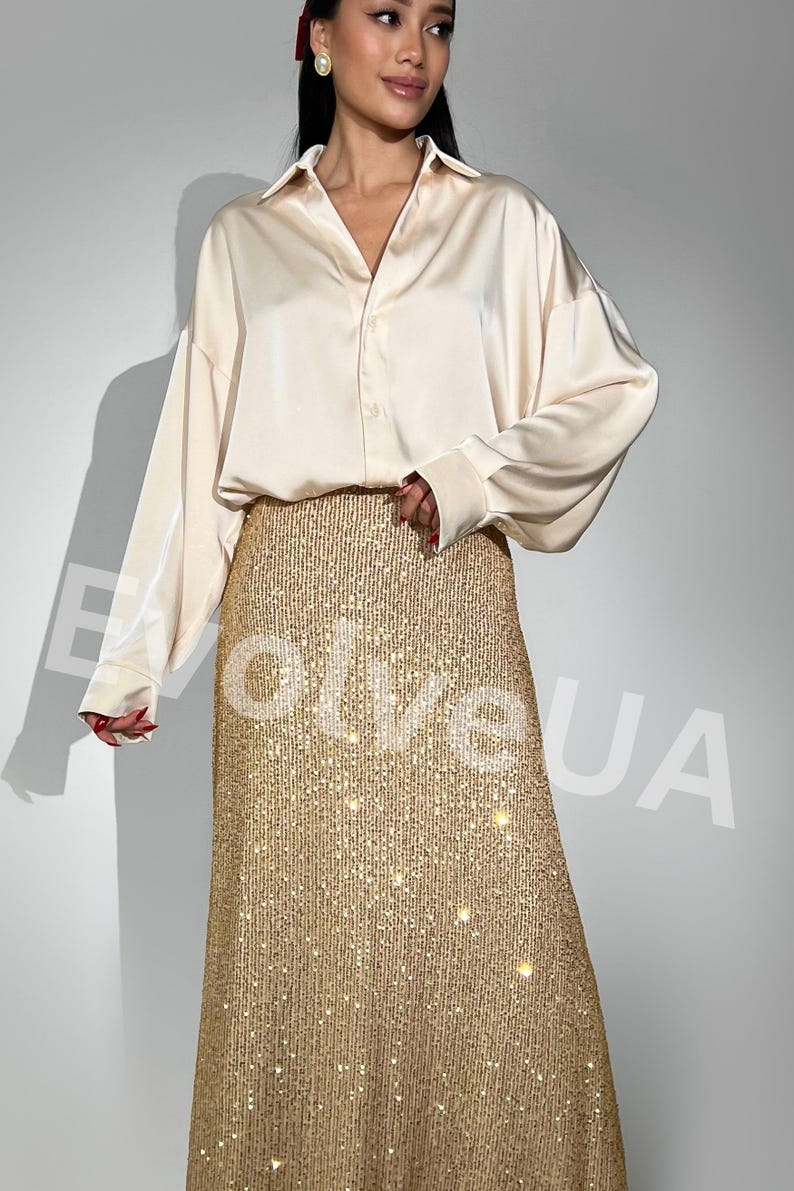May include: A woman is wearing a long, gold sequined skirt with a cream colored satin shirt. The skirt has a subtle shimmer and is a great choice for a special occasion.