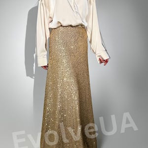 May include: A long, gold sequined skirt with a flowing silhouette. The skirt is paired with a white satin button-down shirt.