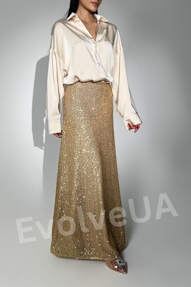 May include: A long, gold sequined skirt with a flowing silhouette. The skirt is paired with a white satin button-down shirt.