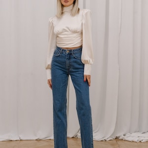 Full-length front view of the ivory open back turtleneck blouse with bishop sleeves, fitted bodice, and smooth satin-finish silk blend fabric, on model styled with blue jeans and black heels; white backdrop.