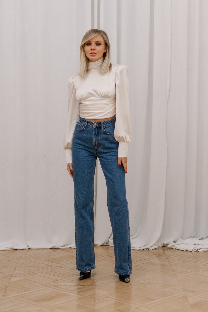 Full-length front view of the ivory open back turtleneck blouse with bishop sleeves, fitted bodice, and smooth satin-finish silk blend fabric, on model styled with blue jeans and black heels; white backdrop.