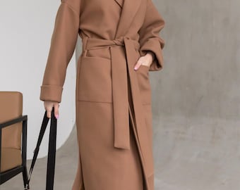 May include: A long, camel-colored coat featuring a wide lapel, long sleeves with cuffs, and a matching fabric tie belt at the waist. The coat includes two large front patch pockets and falls to a mid-calf length, worn over blue denim jeans.
