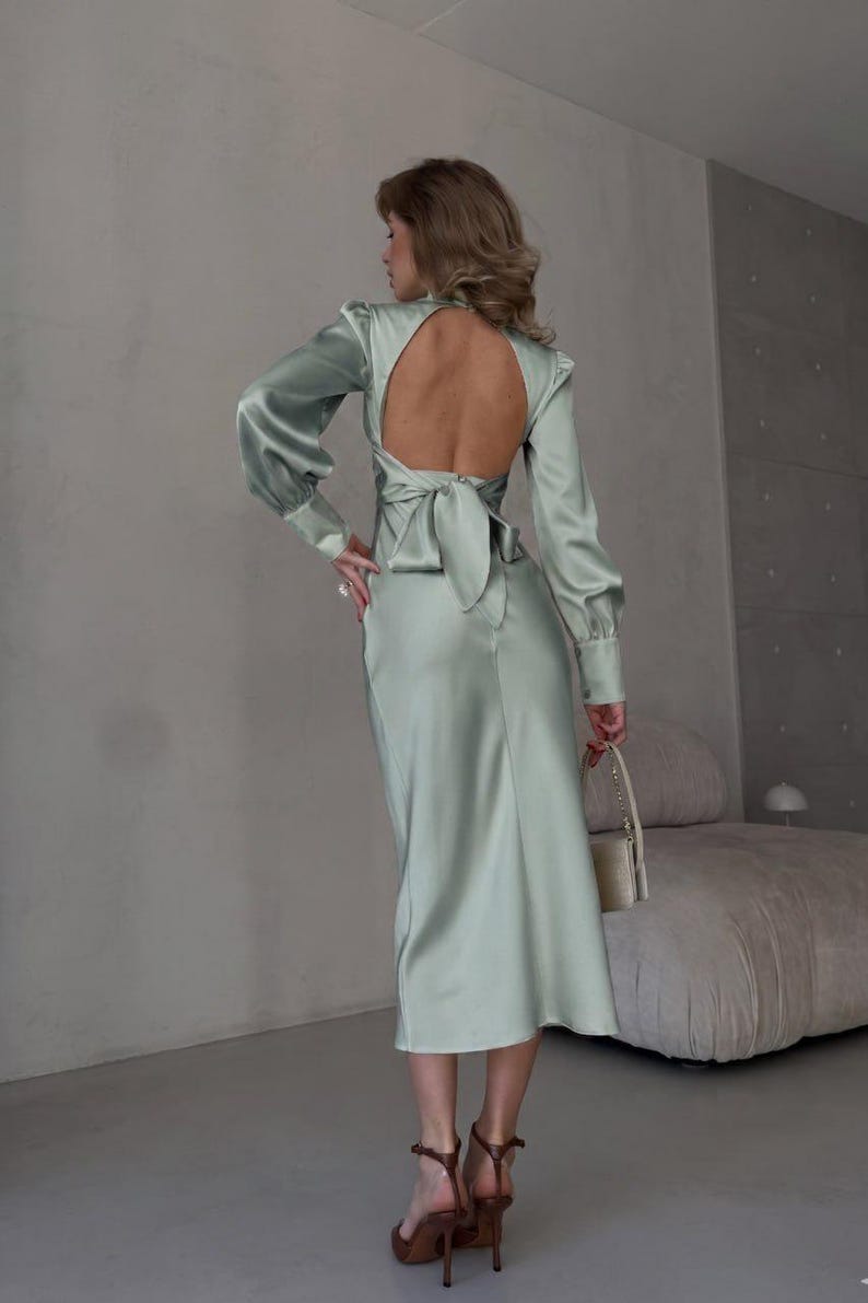 Full-length back view of sage green satin midi dress showing the open back construction and self-tie bow detail at the center back styled with gold heels