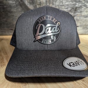 Peut inclure: Un chapeau de camionneur noir et gris avec un patch argenté qui dit "Bientôt papa à nouveau" avec une paire d'empreintes de pas de bébé. Le chapeau a un dos en maille noire et une étiquette blanche "Krafty".