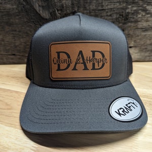 Peut inclure: Un chapeau de camionneur gris avec un patch en cuir marron qui dit "DAD Quinn & Harper". Le chapeau a un dos en maille noire et une visière grise.