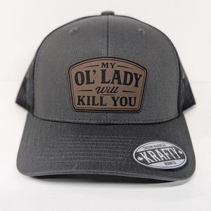 May include: A charcoal grey trucker hat with a black mesh back and a brown rectangular patch on the front. The patch features the black text MY OL' LADY Will KILL YOU. A circular sticker on the brim reads CUSTOM HEADWEAR KRAFTY DESIGN CO.