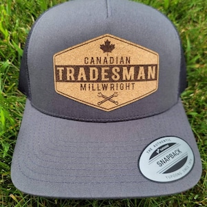 May include: A grey and black trucker hat with a cork patch featuring the text "Canadian Tradesman Millwright" and a maple leaf. The hat has a black mesh back and a white sticker that says "The Authentic Yupoong Snapback Yupoong Since 1974".