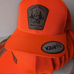 May include: Orange and brown hunting hat with a mesh back. The hat has a patch that says "Swamp Shack Hunt Club Est. 2022". The hat also has a black and white label that says "Krafty Design Co."