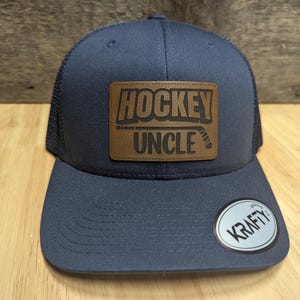May include: A navy blue trucker hat with a brown leather patch that says "Hockey Uncle" in white lettering. The hat has a mesh back and a black adjustable strap.