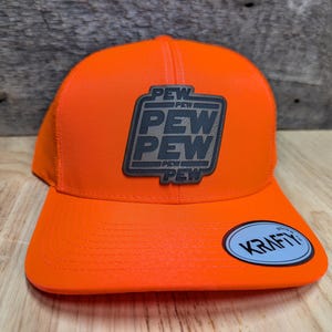 May include: Orange and brown trucker hat with a brown leather patch that says "PEW PEW PEW" and a white label that says "KRAFTY" on the brim.