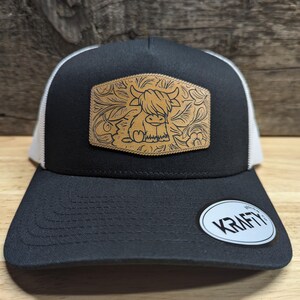 May include: Black trucker hat with a white mesh back. The hat features a brown leather patch with a floral design and a cartoon Highland cow.