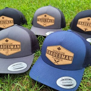 May include: Five different trucker hats with a black mesh back. Each hat has a brown, rectangular patch with the text "Canadian Tradesman" and the name of a trade. The trades are Carpenter, Millwright, Electrician, Sheet Metal Worker, and Plumber.