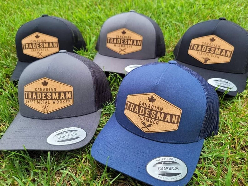 May include: Five different trucker hats with a black mesh back. Each hat has a brown, rectangular patch with the text "Canadian Tradesman" and the name of a trade. The trades are Carpenter, Millwright, Electrician, Sheet Metal Worker, and Plumber.