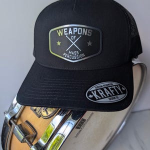 Peut inclure: Casquette de camionneur noire avec un dos en filet et une visière incurvée. L'avant présente un écusson avec le texte "WEAPONS OF MASS PERCUSSION" et des baguettes croisées. Une petite étiquette indique "KRAFTY DESIGN CO."