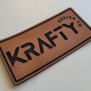 May include: A brown leather patch with black stitching and the text "Krafty Design Co." in black lettering.