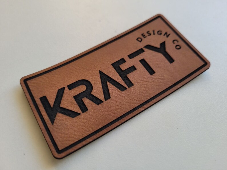May include: A brown leather patch with black stitching and the text "Krafty Design Co." in black lettering.