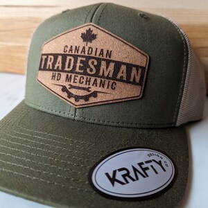 May include: A green and beige trucker hat with a brown leather patch that says "Canadian Tradesman HD Mechanic". The patch features a maple leaf and a wrench and gear design. The hat also has a white oval patch that says "Krafty Design Co."