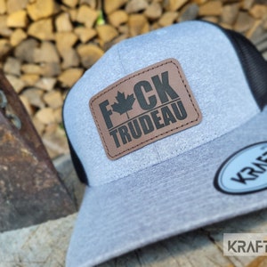 Peut inclure: Un chapeau de camionneur gris et noir avec un patch en cuir brun qui dit "F*CK TRUDEAU" avec une feuille d'érable canadienne. Le chapeau a un dos en maille noire et un devant gris.