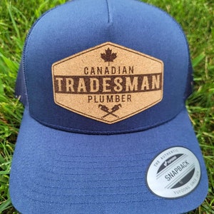 Peut inclure: Un chapeau de camionneur bleu marine avec un patch en liège qui dit "Canadian Tradesman Plumber" avec une feuille d'érable et des clés à molette croisées. Le chapeau a un dos en maille noire et un autocollant blanc "The Authentic Yupoong Snapback".