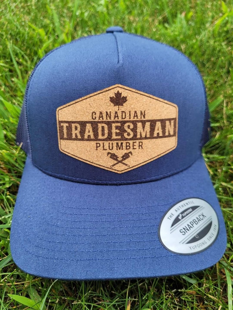May include: A navy blue trucker hat with a cork patch that says "Canadian Tradesman Plumber" with a maple leaf and crossed wrenches. The hat has a black mesh back and a white "The Authentic Yupoong Snapback" sticker.