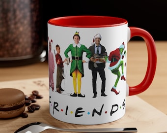 May include: White ceramic mug with a red rim and handle. The mug features a graphic of characters from popular Christmas movies, including Buddy the Elf, The Grinch, and Clark Griswold. The text "FRIENDS" is printed below the characters in a colorful font.