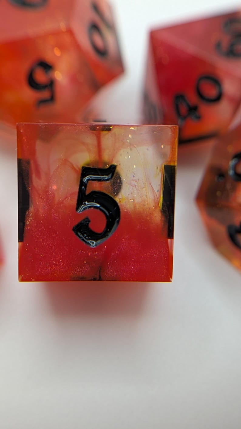 May include: Close-up of a set of handmade dice. The focus is on a square die with the number 5 in black. The die is transparent with red and orange hues and glitter. Other dice are visible in the background, out of focus.
