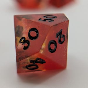 May include: A close-up of a red, multi-sided die with black numbers. The die has a speckled, translucent appearance with gold and black accents. The numbers 20, 30, and 40 are visible. This is a gaming accessory.