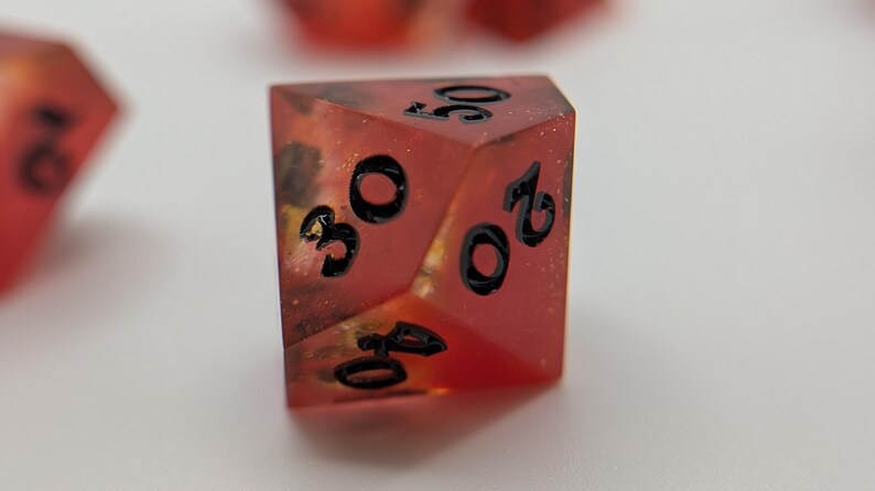 May include: A close-up of a red, multi-sided die with black numbers. The die has a speckled, translucent appearance with gold and black accents. The numbers 20, 30, and 40 are visible. This is a gaming accessory.