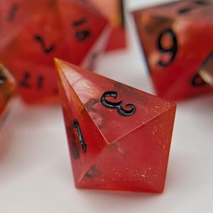 May include: A set of red, multi-sided dice with black numerals. The dice are translucent, with a fiery, speckled interior. One die is in focus, showing the number 3. These dice are likely used for tabletop games.