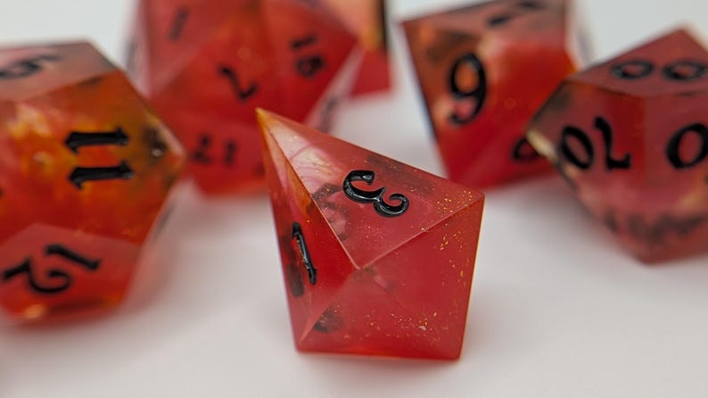 May include: A set of red, multi-sided dice with black numerals. The dice are translucent, with a fiery, speckled interior. One die is in focus, showing the number 3. These dice are likely used for tabletop games.