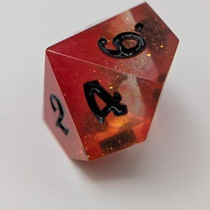 May include: A multi-sided die with a red and orange translucent design, featuring black numerals. The die has a glittery, speckled interior. The numbers 2, 4, and 9 are visible on the die's faces. Suitable for tabletop gaming.