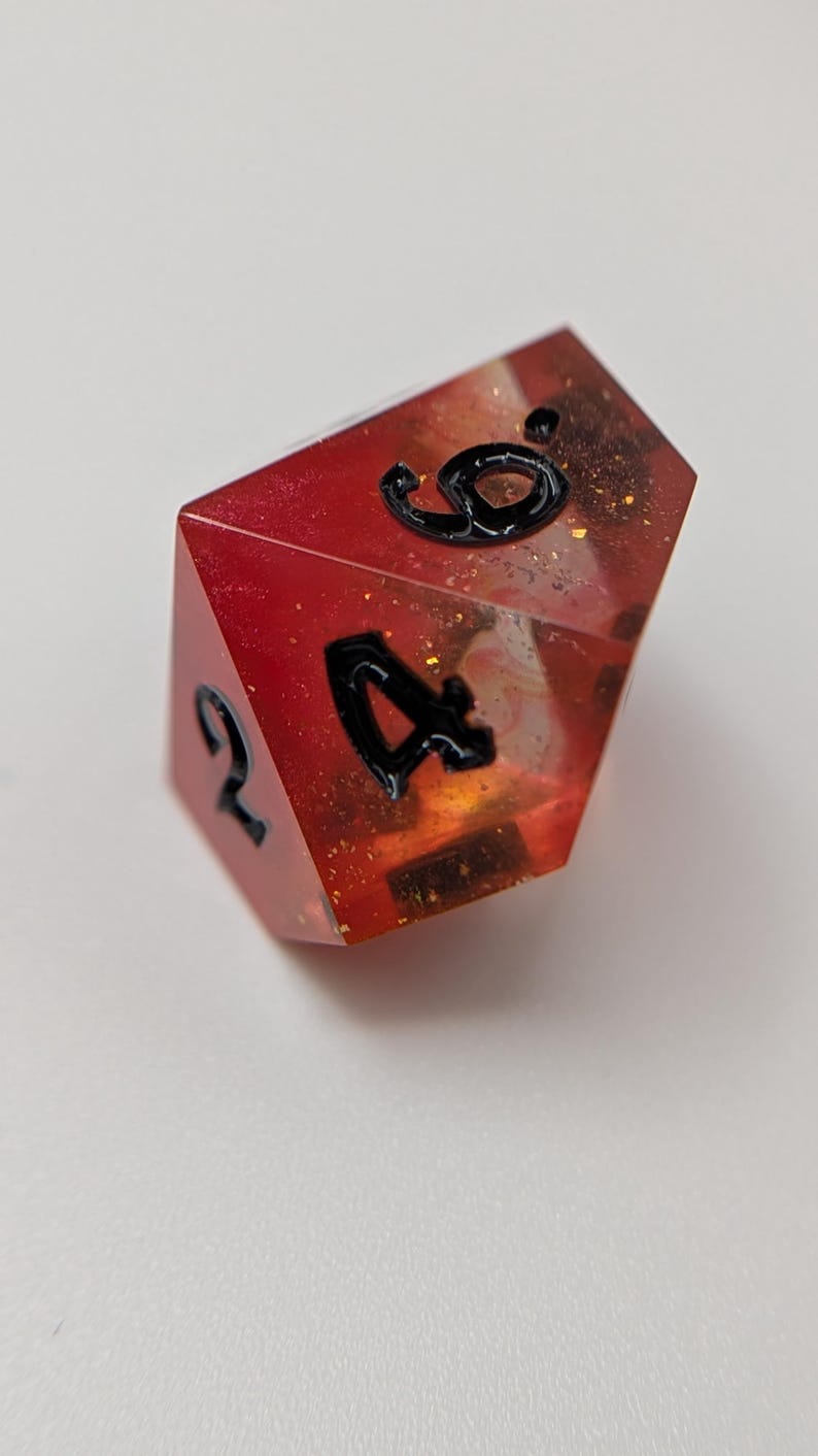 May include: A multi-sided die with a red and orange translucent design, featuring black numerals. The die has a glittery, speckled interior. The numbers 2, 4, and 9 are visible on the die's faces. Suitable for tabletop gaming.