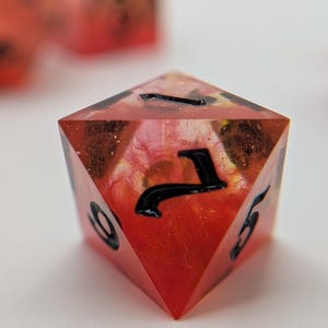May include: A red and orange polyhedral die with black numbering. The die has a translucent appearance, with a swirling pattern of colours inside. Other dice are visible in the background, out of focus.