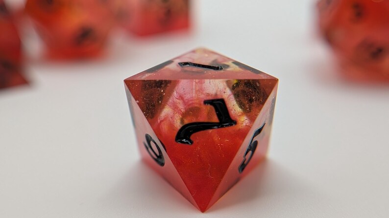 May include: A red and orange polyhedral die with black numbering. The die has a translucent appearance, with a swirling pattern of colours inside. Other dice are visible in the background, out of focus.