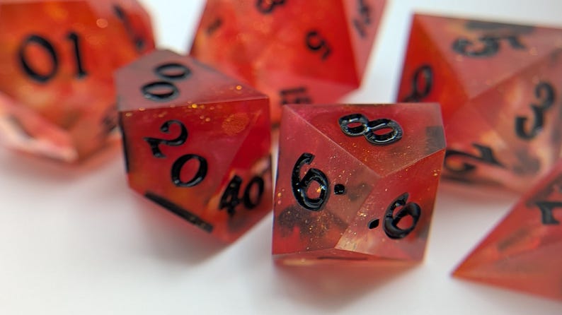 May include: A collection of red polyhedral dice with black numerals. The dice have a translucent quality with gold flecks, designed for role-playing games. The dice are displayed on a white surface.