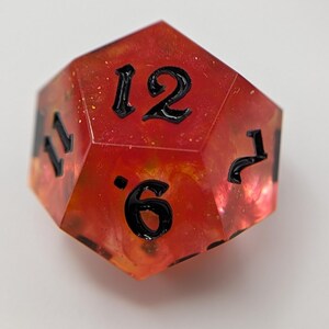 May include: A multi-sided, red and orange resin die with black numbering. The die features the numbers 6, 7, 11, and 12. The die is designed for tabletop gaming.