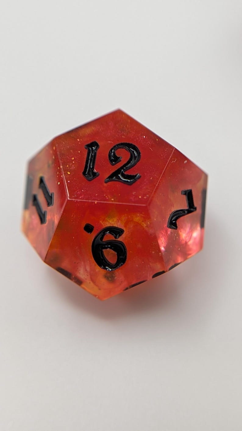 May include: A multi-sided, red and orange resin die with black numbering. The die features the numbers 6, 7, 11, and 12. The die is designed for tabletop gaming.