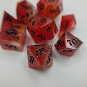 May include: A set of red and black polyhedral dice, with black numerals. The dice are made of a translucent material, with a speckled pattern. The dice are in various shapes, including a d20, d10, d8, d6, and d4, used in tabletop role-playing games.
