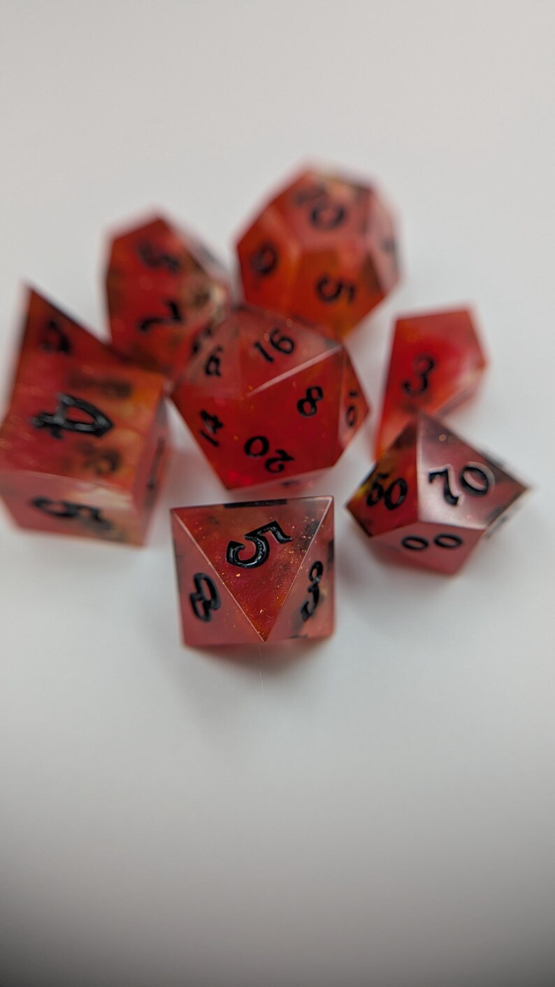 May include: A set of red and black polyhedral dice, with black numerals. The dice are made of a translucent material, with a speckled pattern. The dice are in various shapes, including a d20, d10, d8, d6, and d4, used in tabletop role-playing games.