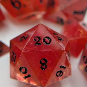 May include: A collection of red polyhedral dice with black numerals. The dice are made from a translucent material, revealing their internal structure. The image features several dice, including a prominent twenty-sided die, perfect for role-playing games.