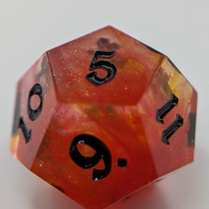 May include: A twelve-sided polyhedral die with a red and orange interior, and black numbers. The die displays the numbers 5, 9, 10, and 11. This unique die is suitable for tabletop gaming.