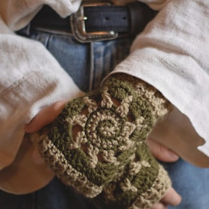 May include: Olive green and tan crocheted fingerless gloves. The gloves have an intricate openwork design and are worn over a white long-sleeved shirt and blue jeans. A handmade accessory.