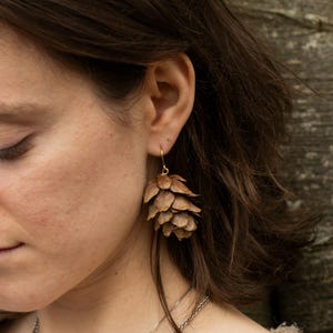 Può includere: Primo piano di una persona che indossa orecchini marroni a forma di cono con ganci color oro. Gli orecchini sono fatti di materiali naturali e pendono dall'orecchio. La persona indossa una collana d'argento e un maglione fantasia.