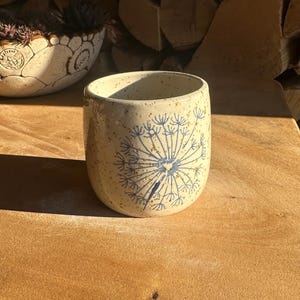 Puede incluir: Una taza de cerámica beige con un diseño de diente de león azul. La taza es cilíndrica con un acabado moteado. Un segundo cuenco decorativo de cerámica con un diseño floral está en el fondo. Los objetos están sobre una superficie de madera.